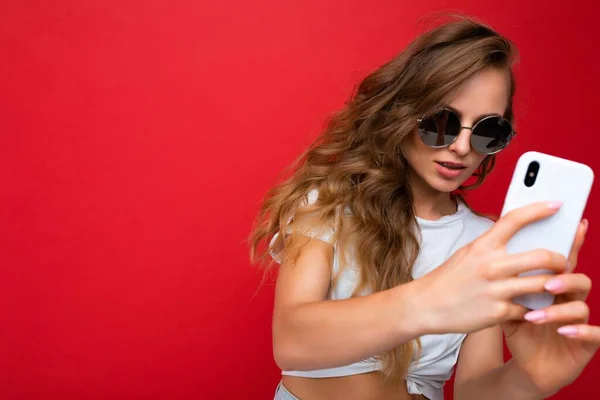 Incrível bela jovem segurando telefone celular tirando foto selfie usando câmera smartphone vestindo roupa elegante todos os dias isolado sobre fundo de parede colorido olhando para a tela do dispositivo — Fotografia de Stock