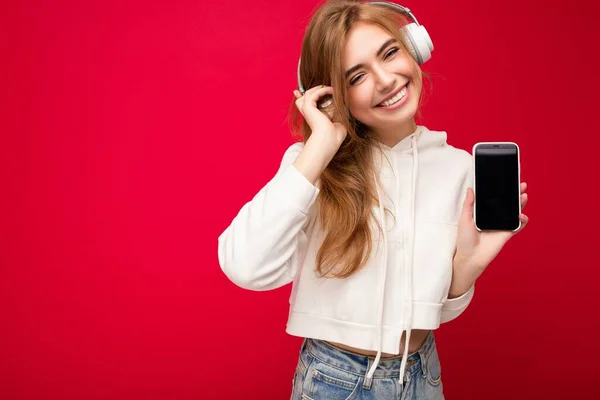 멋진 미소를 짓고 있는 아름다운 젊은 여성의 사진 이 배경 벽에 고립되어 있고, 음악을 듣고 있는 흰색 블루투스 헤드폰을 쓰고 있는 휴대 전화를 사용하고 있다. — 스톡 사진