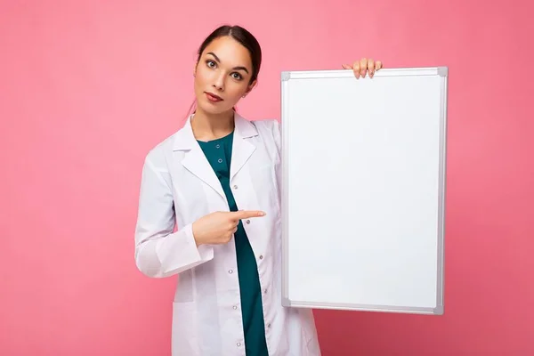Giovane di bella donna brunetta felice indossa cappotto bianco medico tenendo bordo magnetico bianco per mockup isolato su sfondo rosa con spazio vuoto — Foto Stock