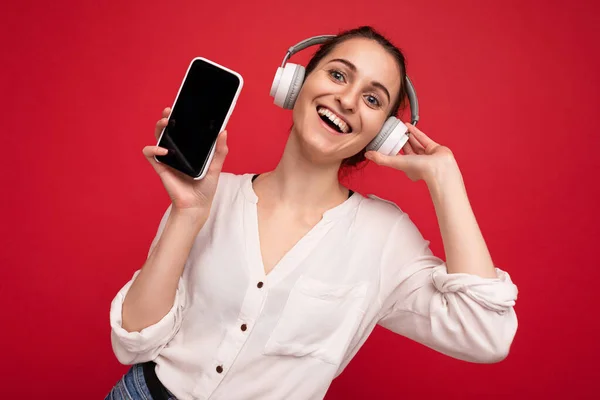 Beautiful happy smiling young woman wearing stylish casual outfit isolated on background wall holding and showing mobile phone with empty display for mockup wearing white bluetooth headphones — Stock Photo, Image