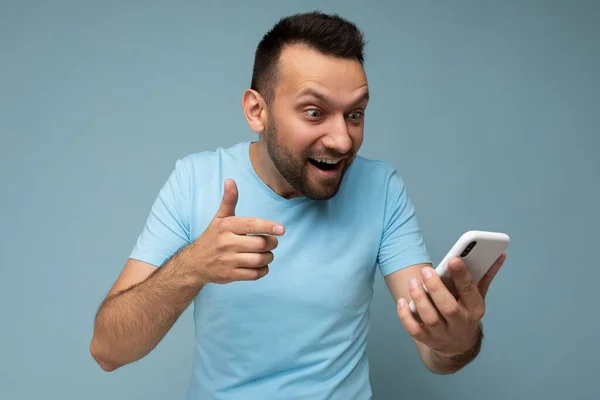 Удивлен красивый молодой бородатый брюнет мужчина в повседневной синей футболке изолированы на синем фоне холдинг и с помощью мобильной связи в Интернете, глядя на gadjet дисплей — стоковое фото