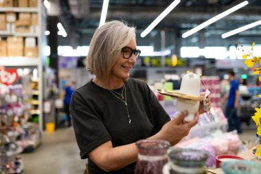 Gri saçlı ve gözlüklü bir kadın, hareketli müşteriler ve parlak vitrinlerle çevrili işlek bir mağazada dekoratif bir parçayı incelerken gülümsüyor..