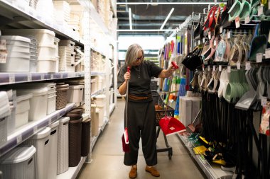 Bir kadın parlak bir ev eşyaları mağazasında temizlik aletlerini inceliyor. Elinde kırmızı bir faraş tutuyor, dikkatle diğer eşyalarla karşılaştırıyor..