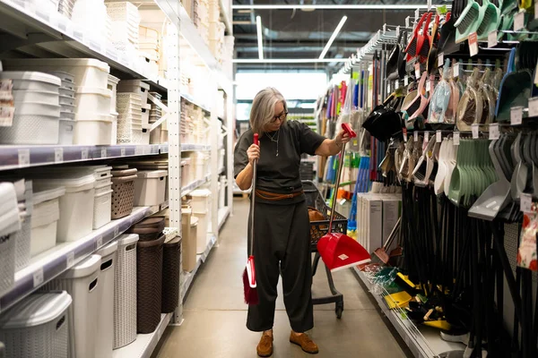 Bir kadın parlak bir ev eşyaları mağazasında temizlik aletlerini inceliyor. Elinde kırmızı bir faraş tutuyor, dikkatle diğer eşyalarla karşılaştırıyor..