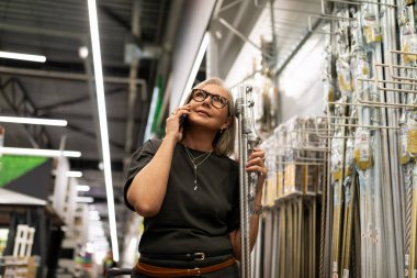 Gözlüklü bir kadın, bir mağazada çeşitli ev geliştirme ürünlerine bakarken telefonda konuşuyor. Düşünceli ve alışverişiyle meşgul görünüyor..