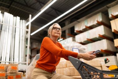 Dolu bir depoda, turuncu kazaklı bir kadın alışveriş arabasında otururken akıllı telefonunu kullanıyor. Kutu yığınları, devam etmekte olan bir alışveriş gezisini gösteren yoğun bir arkaplan oluşturur.