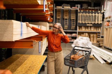 Bir kadın, ev dekorasyonu mağazasında ahşap kalasları inceliyor. Turuncu bir kazak ve bej pantolon giyiyor, kendi seçimiyle ilgileniyor. Yakınlarda bir alışveriş arabası var, araç gereç ve malzeme taşıyor..