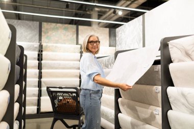 Bir kadın, çeşitli tasarımlarla dolu bir ev dekoru mağazasında büyük bir duvar kağıdı örneğini inceliyor. Rafların yanında duruyor ve seçimine odaklanmış görünüyor..