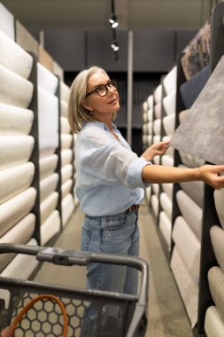 Gözlüklü bir kadın, bir dekorasyon dükkanında duvar kağıdı rulolarını karıştırıyor. Farklı desenleri ve dokuları incelerken odaklanmış ve meşgul görünüyor..