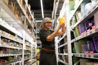 Gözlük takan gülümseyen bir kadın, iyi organize edilmiş bir mağazanın rafından sarı bir kap seçiyor. Etrafını parlak renkler sarmış, çeşitli mutfak ve ev eşyaları sergiliyor..