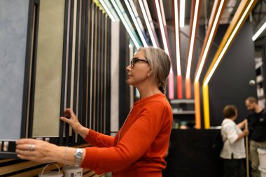 Bir kadın bir tasarım sergisinde farklı duvar malzemelerini inceliyor. Kabin canlı ışıklandırma ve çeşitli dokularla dolu, yenilikçi tasarım eğilimleri sergiliyor..