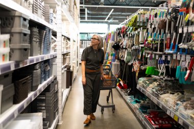 Bir kadın, parlak bir mağazadaki çeşitli ev organizasyonlarıyla dolu rafları karıştırıyor. Bir alışveriş arabası tutuyor ve ürünleri dikkatlice inceliyor..