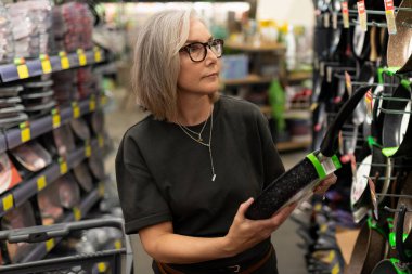 Bir kadın iyi stoklanmış bir mutfak malzemesi dükkanında alışveriş yaparken bir kızartma tavasını inceler. Etrafı çeşitli tencere ve tavalarla çevrili..