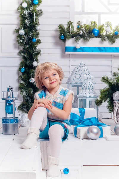 Smiling little blonde sweet girl sitting on the veranda surrounded by white Christmas balls and Christmas tree — Stock Photo, Image