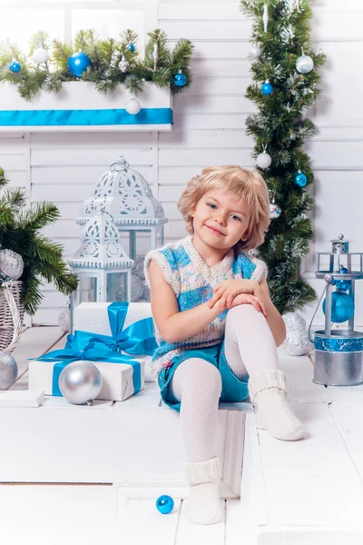 Lächelndes kleines blondes süßes Mädchen sitzt auf der Veranda, umgeben von weißen Weihnachtskugeln und Weihnachtsbaum — Stockfoto