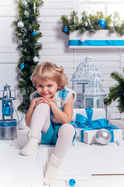 Smiling little blonde sweet girl sitting on the veranda surrounded by white Christmas balls and Christmas tree — Zdjęcie stockowe