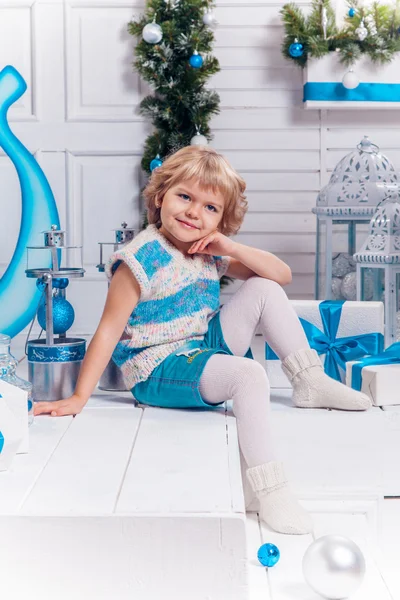 Lächelndes kleines blondes süßes Mädchen sitzt auf der Veranda, umgeben von weißen Weihnachtskugeln und Weihnachtsbaum — Stockfoto