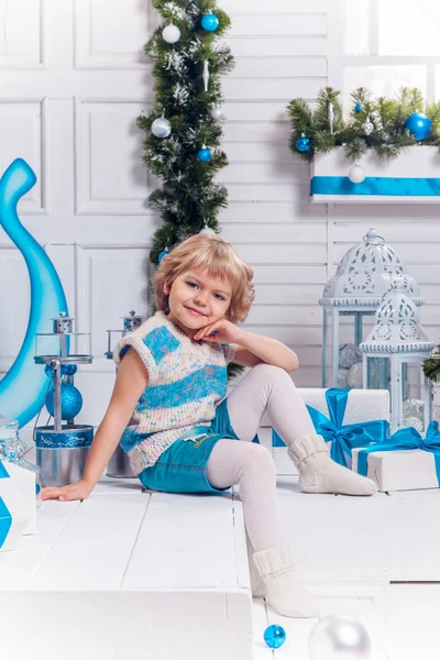 Улыбающаяся маленькая блондинка милая девочка сидит на веранде в окружении белых рождественских шаров и рождественской елки — стоковое фото