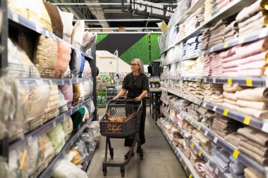 Bir kadın bir alışveriş arabasını bir ev eşyaları mağazasının koridoruna itiyor, çeşitli renkli yastıkları ve kumaşları geziyor, alışveriş deneyiminin tadını çıkarıyor..