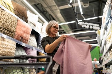 Gözlüklü bir kadın, ev eşyaları mağazasındaki renkli tekstil ürünleri arasında yumuşak pembe bir battaniyeyi inceliyor. Alışveriş yaparken alışverişini düşünürken düşünceli görünüyor..