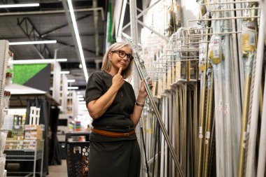 Bir kadın mağaza koridorunda durup çeşitli materyalleri inceliyor. Elinde uzun bir şey var ve seçeneklerini değerlendiriyor gibi görünüyor..