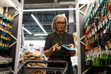 Son sınıf öğrencisi bir kadın, elinde ambalaj tutarken bir perakende mağazasının koridorunda bahçe aletlerini inceliyor. Bir alışveriş arabası var ve seçimine odaklanmış görünüyor..