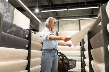 Kısa gri saçlı bir kadın, çeşitli doku ve renklerle dolu bir koridorda dururken tasarım malzemelerini inceliyor. Elinde kağıt tutarken odaklanmış görünüyor..