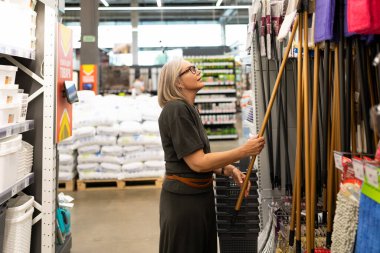 Gri saçlı bir kadın, yoğun bir alışveriş gününde, çeşitli ev eşyaları ve malzemelerle çevrili bir mağaza koridorunda temizlik aletlerini inceliyor..