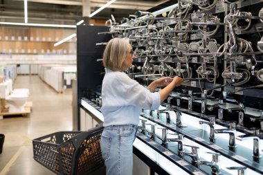 Bir kadın, bir ev geliştirme mağazasında çeşitli muslukları kontrol eder. Serginin yanında duruyor, elinde parlak bir muslukla alışveriş arabasına bir şeyler yerleştiriyor..