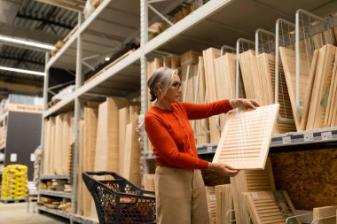 Bir kadın, bir ev geliştirme mağazasındaki ahşap panellere yakından bakıyor. Malzemelerle dolu bir arabanın yanında duruyor. Yoğun koridorda çeşitli malzemelerle dolu tercihlerini düşünüyor..