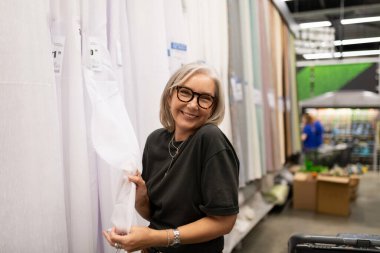 Neşeli bir kadın el sanatları mağazasında dururken bir rulo kumaş tutuyor. Parlak ışık, raflardaki çeşitli materyaller arasından seçimleri incelerken içeri süzülüyor.