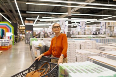 Gözlüklü bir kadın, içi ev eşyalarıyla dolu bir dükkanda alışveriş arabasını iterken mutlu görünür. Şık bir kıyafet giyip seçeneklerini değerlendiriyor..