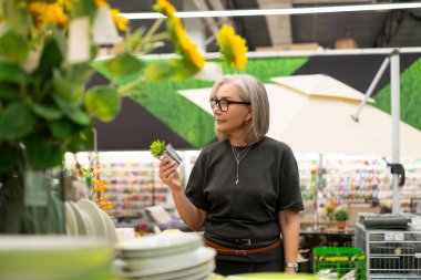 Gri saçlı bir kadın renkli çiçekler ve bahçe aletleriyle dolu bir bahçe merkezine bakıyor. Seçeneklerini göz önünde bulundurarak küçük bir bitki tutuyor..