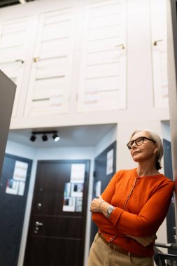 Gümüş saçlı olgun bir kadın şık bir koridorda duruyor, turuncu bir kazak giyiyor. Duvara yaslanırken sakin ve düşünceli görünüyor. Çağdaş uzayın atmosferinin tadını çıkarıyor..