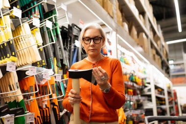 Gözlüklü bir kadın, çeşitli araç gereç ve gereçlerle dolu bir nalbur reyonunda dikilirken baltayı yakından inceliyor. Düşünceli ve odaklanmış görünüyor..