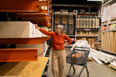 Bir kadın ev dekorasyonu mağazasında duruyor, raftaki tahta tahtalara yakından bakıyor. Kendi projesi için içi malzeme dolu bir alışveriş arabasını itiyor..