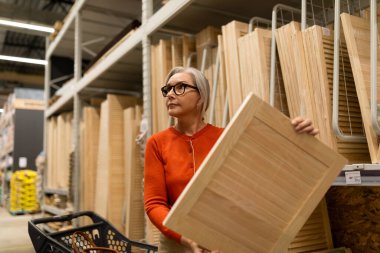 Gri saçlı bir kadın, alışveriş arabasını iterken bir mağazada ahşap bir paneli inceliyor. Bir proje için malzeme seçerken düşünceli ve odaklanmış görünüyor..