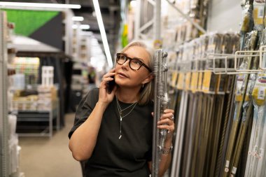 Günlük kıyafet giymiş bir kadın, bir mağazadan alışveriş yaparken telefonla konuşuyor. Bir elinde metal bir nesne tutarken düşünceli görünüyor. Parlak mağaza ışıkları koridoru aydınlatıyor.