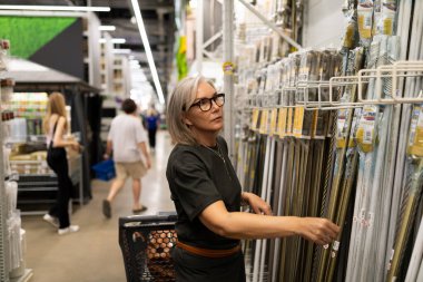 Gri saçlı ve gözlüklü yaşlı bir kadın, bir ev geliştirme mağazasında alet ve malzeme sergisinden eşyalar seçiyor. Diğer müşteriler arka planda görülebilir..