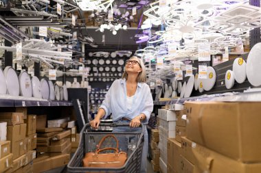 Gözlüklü bir kadın, bir alışveriş arabasını mağazadan geçirirken gülümsüyor. Etrafı çeşitli ışıklandırma seçenekleri ve kutularla çevrili, alışveriş deneyiminin tadını çıkarıyor..