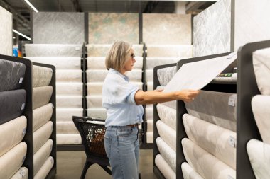 Bir kadın geniş bir yatak mağazasında duruyor ve çeşitli seçeneklere bakıyor. Yanında bir alışveriş arabası varken bir şilte numunesi tutuyor. Bu da yeni yatak aradığını gösteriyor..