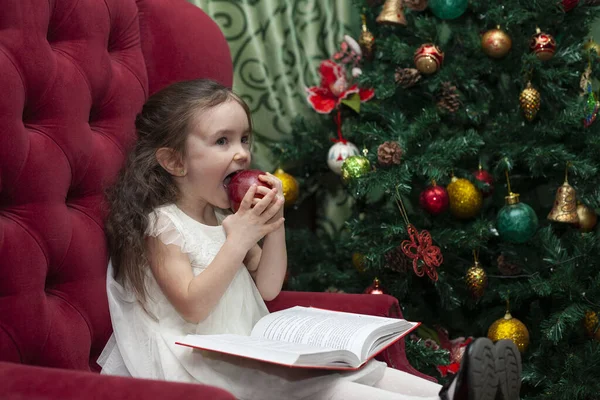 Маленька Дівчинка Читає Книжку Кріслі Біля Ялинки Дитина Їсть Яблуко — стокове фото