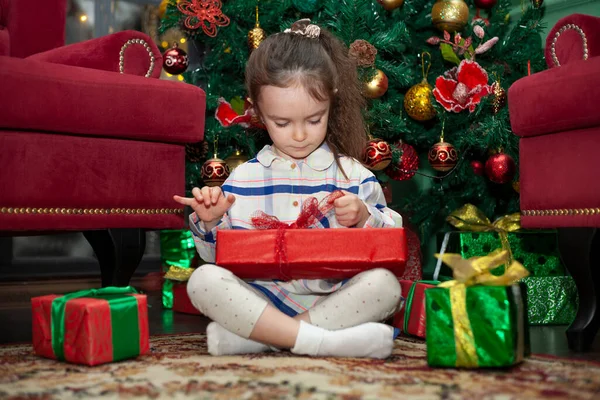 A little girl opens gifts near the Christmas tree. New Year\'s morning. Child and New Year\'s gifts