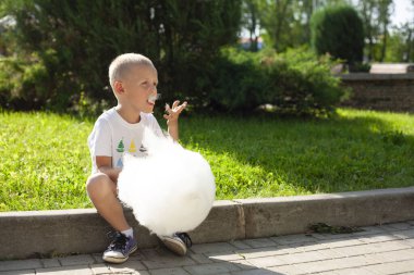 Küçük çocuk parkta kaldırımda oturuyor ve büyük beyaz pamuk şeker yiyor.
