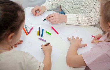 Genç bir anaokulu öğretmeni çocuklarla resim çizer. Anne ve kızları masada kalemle resim çizerler..