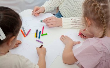 Genç bir anaokulu öğretmeni çocuklarla resim çizer. Anne ve kızları masada kalemle resim çizerler..