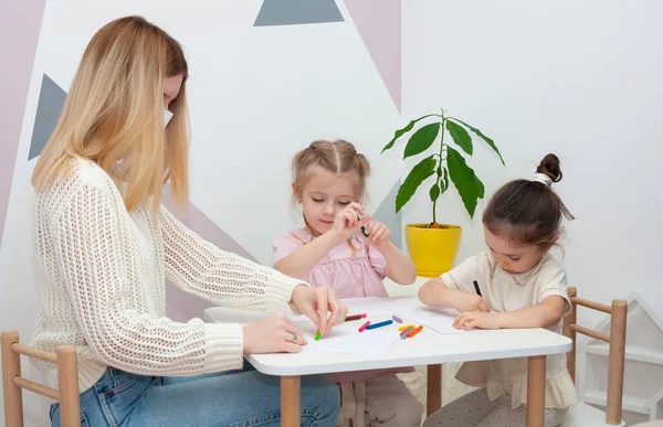 A young kindergarten teacher draws with children. Mom and daughters draw with pencils at the table.
