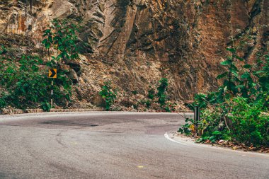 Keskin kıvrımlı dağ yolu yeşil bitkilerle kayalık bir yamaçtan geçiyor, uyarı işareti ve doğal açık hava manzarası..