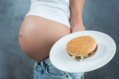 Close up of a cute pregnant belly and junk food. Hamburger and p