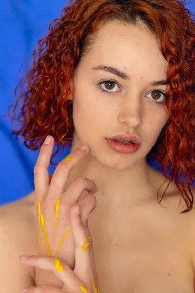 Redhead Curly Woman Hands Yellow Paint Looks Camera Blue Background — Stock Photo, Image